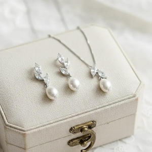 Puede incluir: Un conjunto de joyas con collar y pendientes con dise&ntilde;o de hojas y una perla colgante. El conjunto se exhibe en un joyero crema con cierre dorado. Las joyas son de color plateado.