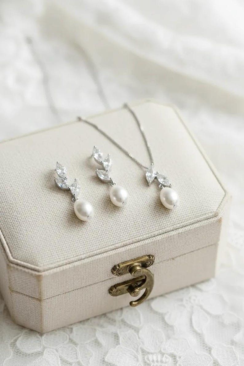 Puede incluir: Un conjunto de joyas con collar y pendientes con dise&ntilde;o de hojas y una perla colgante. El conjunto se exhibe en un joyero crema con cierre dorado. Las joyas son de color plateado.