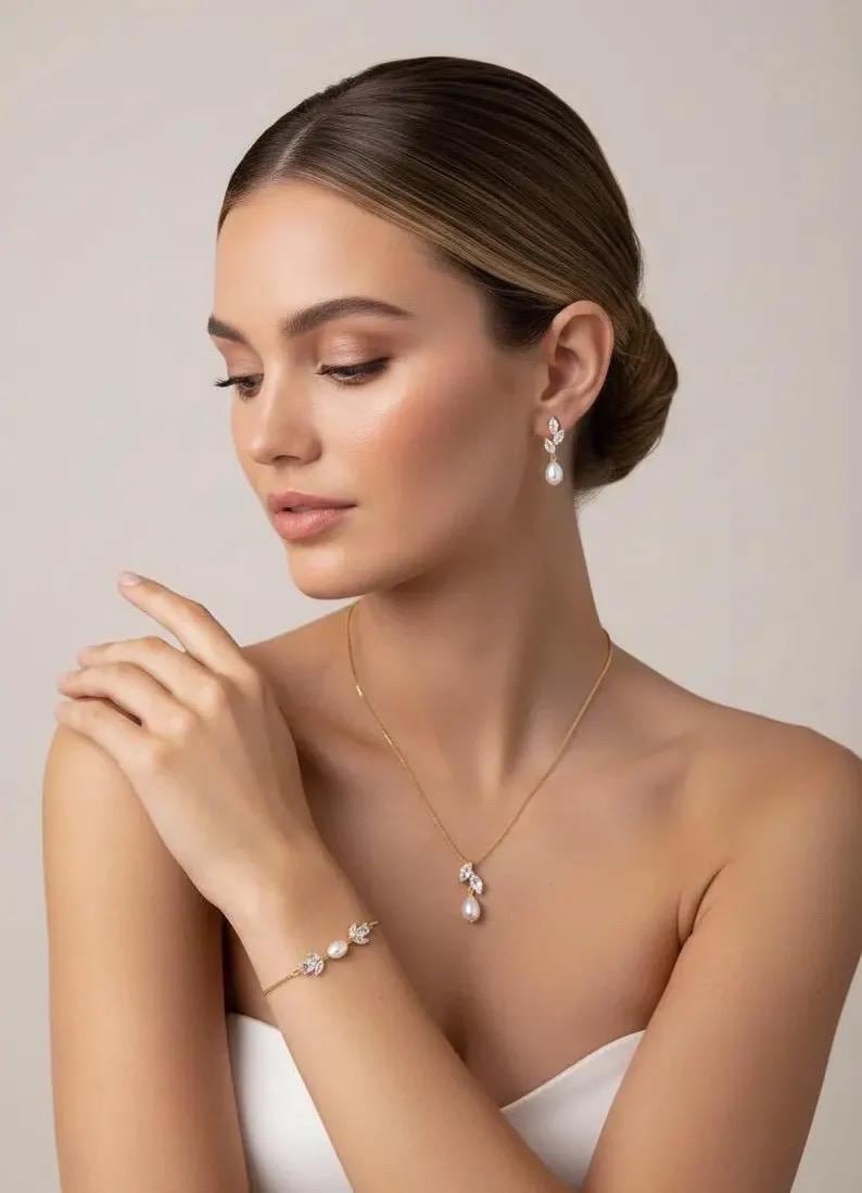Puede incluir: Elegante conjunto de joyas con collar, pulsera y pendientes dorados. El collar y los pendientes tienen un dise&ntilde;o de hojas con una perla colgante. La pulsera tiene perlas y detalles de hojas. Las joyas est&aacute;n sobre un fondo neutro.