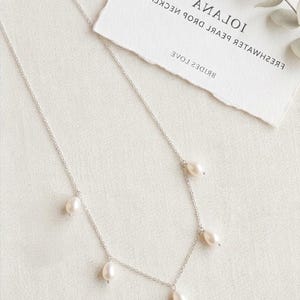 May include: A delicate silver necklace featuring five freshwater pearl drops. The necklace is displayed on a white surface with a tag that reads "IOIANA FRESHWATER PEARL DROP NECKLACE". The Bridal Star logo is visible at the bottom.