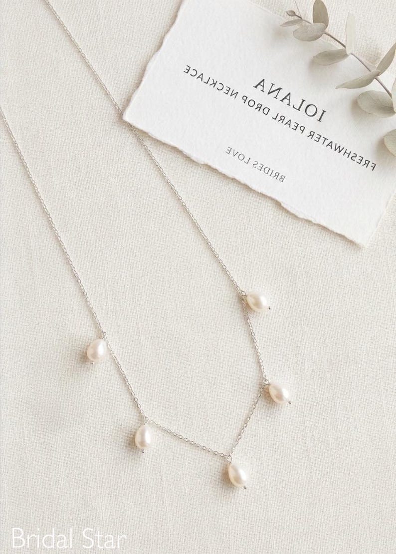 May include: A delicate silver necklace featuring five freshwater pearl drops. The necklace is displayed on a white surface with a tag that reads "IOIANA FRESHWATER PEARL DROP NECKLACE". The Bridal Star logo is visible at the bottom.