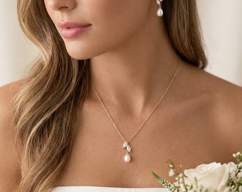 May include: A gold-toned necklace and matching drop earrings set featuring a delicate chain, two marquise-cut clear stones arranged like leaves, and a single white teardrop pearl pendant. The jewelry set is worn with a white strapless garment.