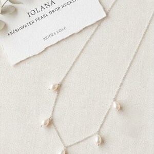 May include: A delicate silver necklace with freshwater pearl drops. The necklace is displayed on a white surface next to a card that reads "IOLANA FRESHWATER PEARL DROP NECKLACE BRIDES LOVE". The necklace has five pear-shaped pearls.