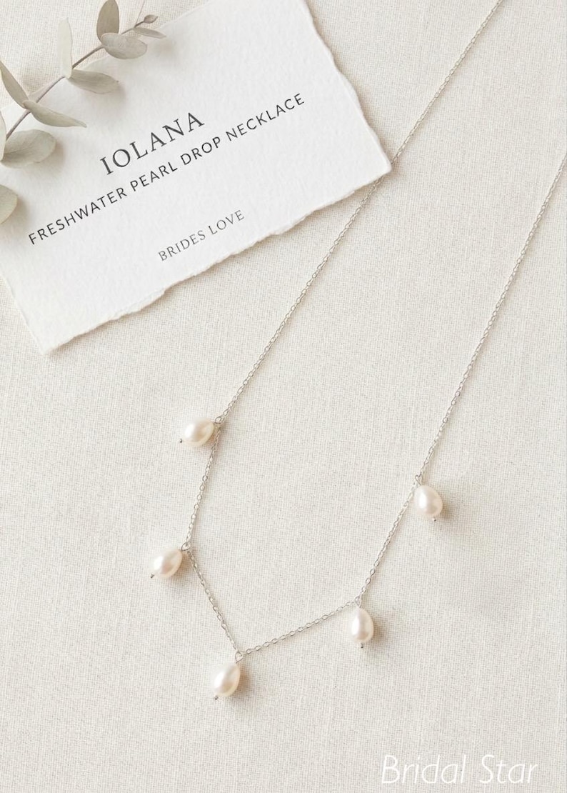 May include: A delicate silver necklace with freshwater pearl drops. The necklace is displayed on a white surface next to a card that reads "IOLANA FRESHWATER PEARL DROP NECKLACE BRIDES LOVE". The necklace has five pear-shaped pearls.