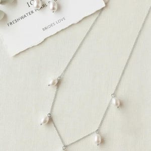 May include: A silver necklace with freshwater pearls and matching earrings. The necklace has a delicate chain with five pearls. The earrings have a pearl drop and a small cluster of clear crystals. The text on the card reads "IC AN FRESHWATER PEARL DROP NECKLACE BRIDES LOVE".