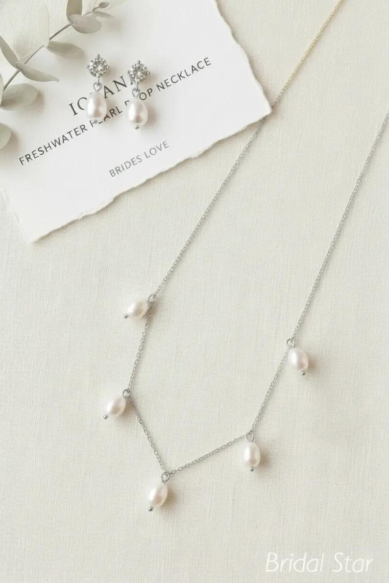 May include: A silver necklace with freshwater pearls and matching earrings. The necklace has a delicate chain with five pearls. The earrings have a pearl drop and a small cluster of clear crystals. The text on the card reads "IC AN FRESHWATER PEARL DROP NECKLACE BRIDES LOVE".