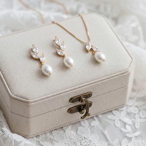 Puede incluir: Un conjunto de joyas dorado con un collar y pendientes, cada uno adornado con cristales transparentes y una perla en forma de l&aacute;grima. El conjunto se exhibe sobre una caja de joyer&iacute;a beige con un cierre de lat&oacute;n, sobre encaje blanco.
