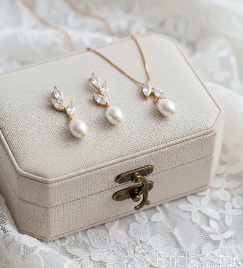 Puede incluir: Un conjunto de joyas dorado con un collar y pendientes, cada uno adornado con cristales transparentes y una perla en forma de l&aacute;grima. El conjunto se exhibe sobre una caja de joyer&iacute;a beige con un cierre de lat&oacute;n, sobre encaje blanco.