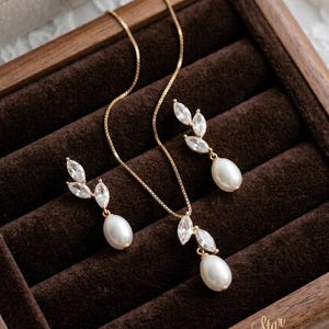 Puede incluir: Un conjunto de joyas dorado, que incluye un collar y pendientes a juego, presentado en una caja de madera. El conjunto presenta un colgante y pendientes con un dise&ntilde;o de hojas de cristales transparentes y una perla en forma de l&aacute;grima. La caja est&aacute; forrada con terciopelo marr&oacute;n.