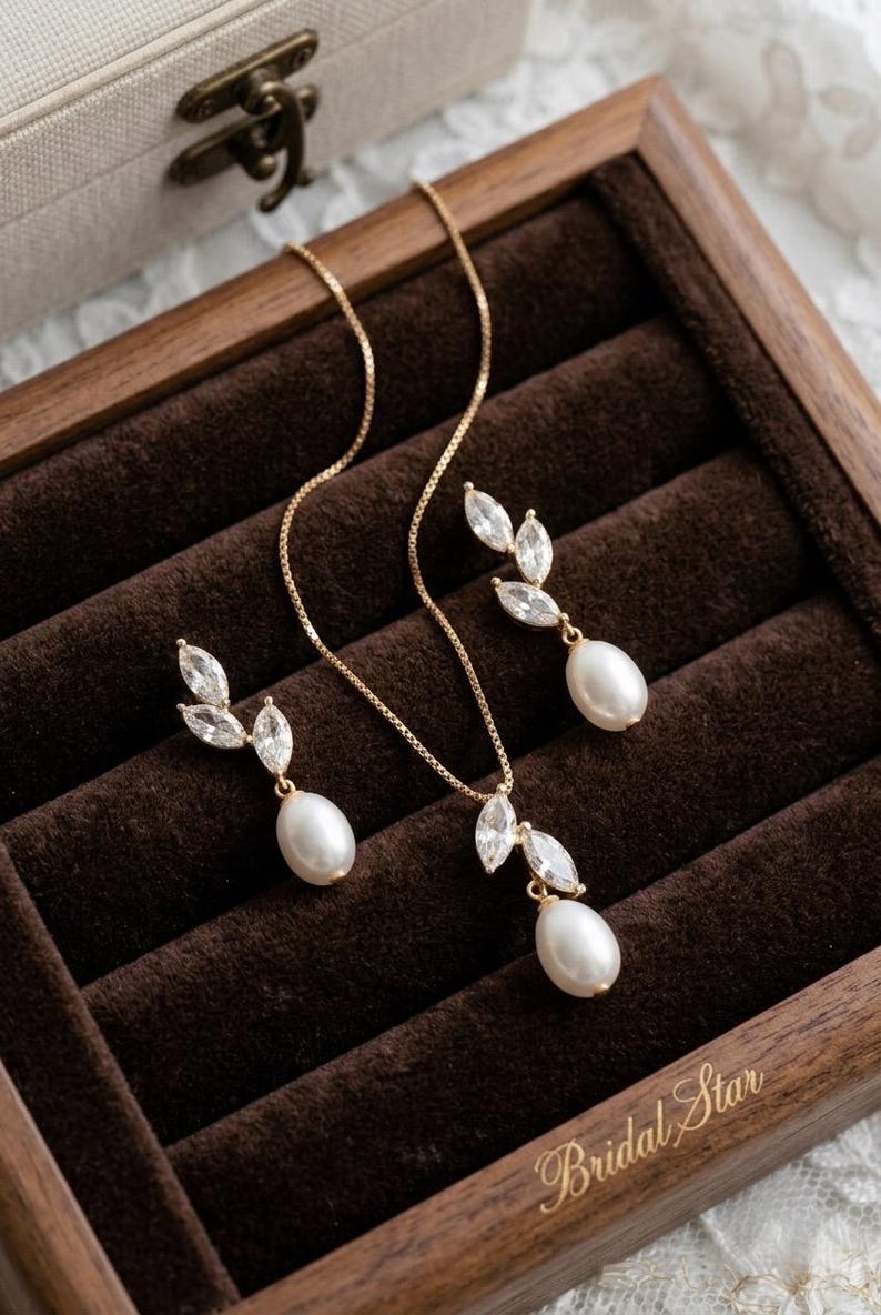 Puede incluir: Un conjunto de joyas dorado, que incluye un collar y pendientes a juego, presentado en una caja de madera. El conjunto presenta un colgante y pendientes con un dise&ntilde;o de hojas de cristales transparentes y una perla en forma de l&aacute;grima. La caja est&aacute; forrada con terciopelo marr&oacute;n.