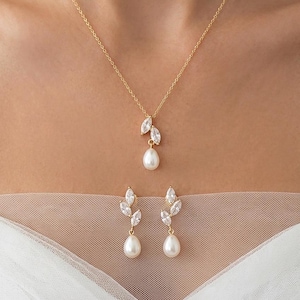 Könnte beinhalten: Ein goldfarbenes Schmuckset bestehend aus einer Halskette und passenden Ohrhängern. Jedes Stück zeigt zwei klare, marquise-geschliffene Kristalle in Blattoptik über einer einzelnen weißen, tropfenförmigen Perle an einer goldenen Kette.