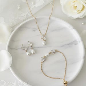 Puede incluir: Un conjunto de joyas doradas que incluye un collar, pendientes y pulsera. Cada pieza tiene un dise&ntilde;o en forma de hoja con cristales transparentes y una perla grande en forma de l&aacute;grima. El conjunto se muestra sobre una superficie blanca con vetas de m&aacute;rmol.