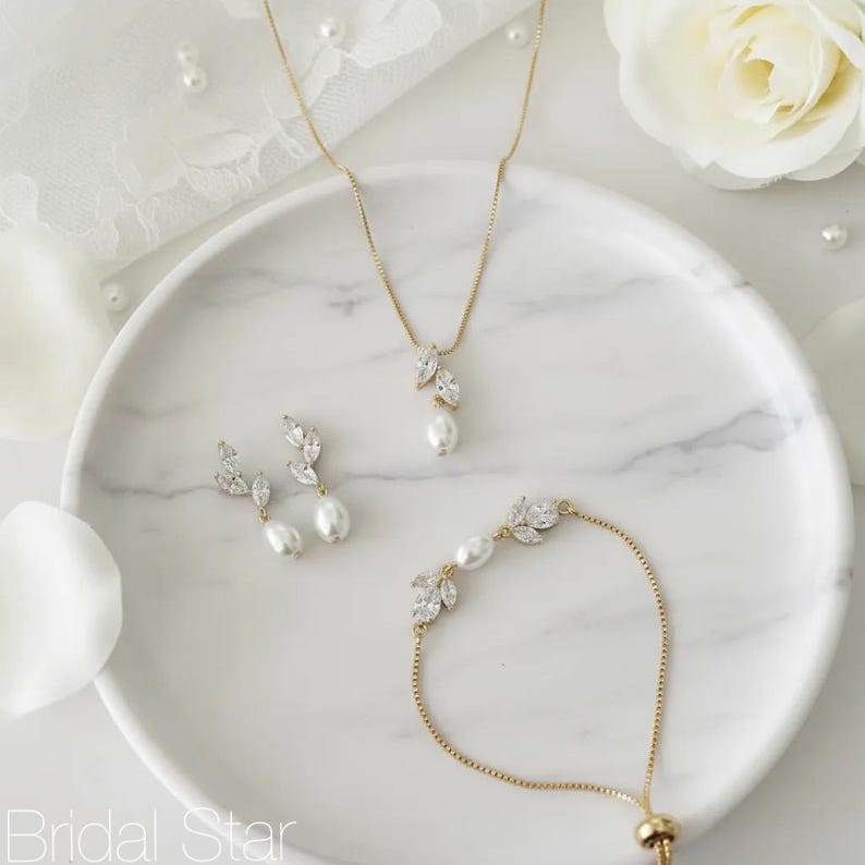 Puede incluir: Un conjunto de joyas doradas que incluye un collar, pendientes y pulsera. Cada pieza tiene un dise&ntilde;o en forma de hoja con cristales transparentes y una perla grande en forma de l&aacute;grima. El conjunto se muestra sobre una superficie blanca con vetas de m&aacute;rmol.