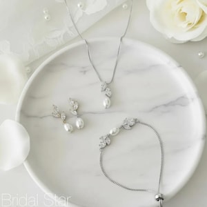 Puede incluir: Un conjunto de joyas de novia que incluye un collar, pendientes y pulsera. El conjunto incluye un collar con un colgante de hoja y perla, pendientes a juego y una pulsera. Las joyas est&aacute;n adornadas con cristales transparentes y perlas blancas, presentadas en un plato de m&aacute;rmol blanco.