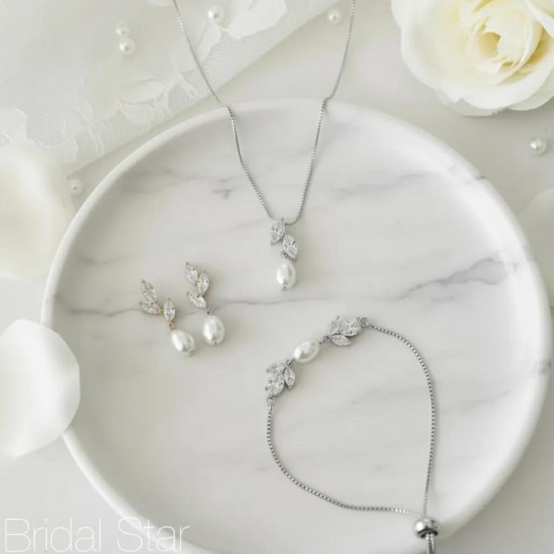 Puede incluir: Un conjunto de joyas de novia que incluye un collar, pendientes y pulsera. El conjunto incluye un collar con un colgante de hoja y perla, pendientes a juego y una pulsera. Las joyas est&aacute;n adornadas con cristales transparentes y perlas blancas, presentadas en un plato de m&aacute;rmol blanco.