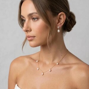 May include: A woman wearing a gold necklace with six white pearls, and matching pearl earrings. The earrings have a small, decorative top. The woman is wearing a white strapless top. Her hair is pulled back in a bun.