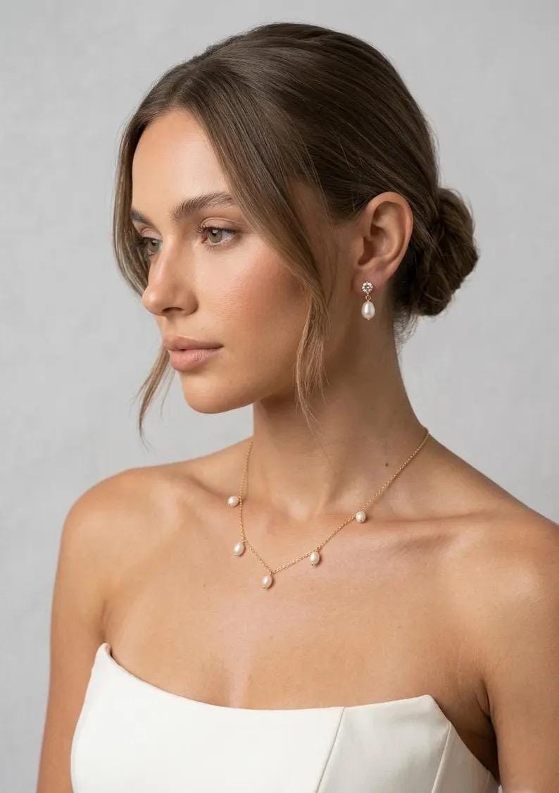 May include: A woman wearing a gold necklace with six white pearls, and matching pearl earrings. The earrings have a small, decorative top. The woman is wearing a white strapless top. Her hair is pulled back in a bun.