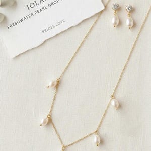 May include: A gold-toned necklace and matching earrings set with freshwater pearl drops. The necklace has multiple pearls suspended from a delicate chain. The earrings feature a pearl drop below a small, sparkling accent. The card reads "IOLANA FRESHWATER PEARL DROP NECKLACE BRIDES LOVE".