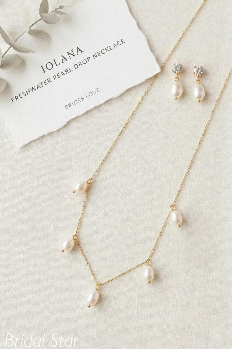 May include: A gold-toned necklace and matching earrings set with freshwater pearl drops. The necklace has multiple pearls suspended from a delicate chain. The earrings feature a pearl drop below a small, sparkling accent. The card reads "IOLANA FRESHWATER PEARL DROP NECKLACE BRIDES LOVE".