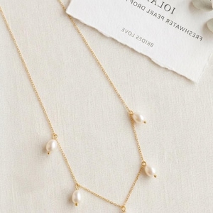 May include: A delicate gold necklace with five freshwater pearls. The necklace is displayed on a white surface with a torn paper tag that reads "Iolana Fresh Water Pearl Drop Necklace" and "Bridal Star".