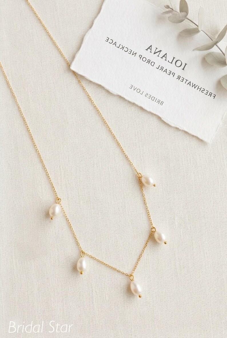 May include: A delicate gold necklace with five freshwater pearls. The necklace is displayed on a white surface with a torn paper tag that reads "Iolana Fresh Water Pearl Drop Necklace" and "Bridal Star".