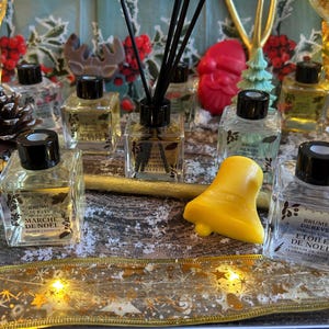 Peut inclure: Plusieurs flacons de parfum en verre transparent avec des bouchons noirs, certains avec des bâtonnets diffuseurs, et une bougie jaune en forme de cloche. Les flacons portent des étiquettes en français, sur un fond décoratif.