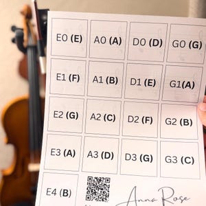 Puede incluir: Una hoja de papel blanca con una cuadrícula de notas musicales y letras, incluyendo E0 (E), A0 (A), D0 (D) y G0 (G). Un violín está ligeramente desenfocado en el fondo. El papel también incluye un código QR y el nombre "Anna Rose".