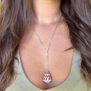 Puede incluir: Un collar de cadena de metal en tono dorado con un colgante estilo gota. El colgante es una concha marina en forma de lágrima con una base blanca y un patrón repetitivo de marcas geométricas en forma de chevrón de color marrón rojizo oscuro.