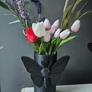 May include: A black vase with a butterfly design, holding a bouquet of flowers. The bouquet includes red roses, white and pink tulips, and purple lavender. The vase has a brick-like texture.