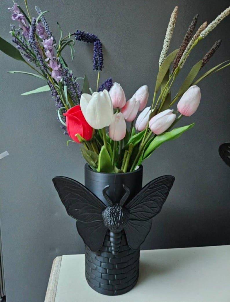 May include: A black vase with a butterfly design, holding a bouquet of flowers. The bouquet includes red roses, white and pink tulips, and purple lavender. The vase has a brick-like texture.