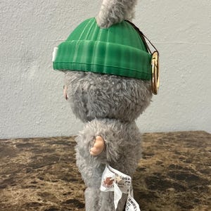 Puede incluir: Un peluche de una peque&ntilde;a criatura peluda con un gorro verde y una oreja gris esponjosa. El juguete tiene una etiqueta adjunta. La criatura es gris con manos y pies rosas. Un anillo dorado est&aacute; unido al gorro.