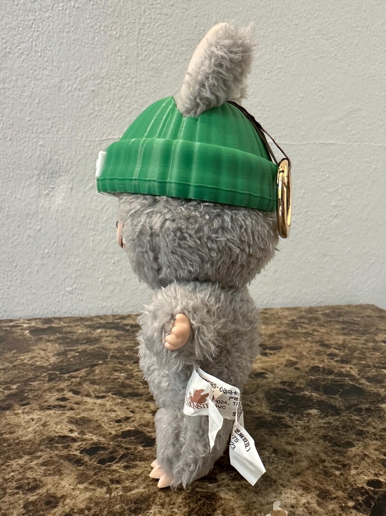 Puede incluir: Un peluche de una peque&ntilde;a criatura peluda con un gorro verde y una oreja gris esponjosa. El juguete tiene una etiqueta adjunta. La criatura es gris con manos y pies rosas. Un anillo dorado est&aacute; unido al gorro.