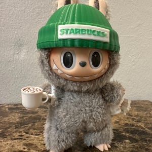 Puede incluir: Una figura de peluche de un personaje de dibujos animados que lleva un gorro verde con "STARBUCKS" impreso. La figura sostiene una peque&ntilde;a taza blanca con frijoles marrones y un papel peque&ntilde;o. La criatura tiene pelaje gris y una cara sonriente.