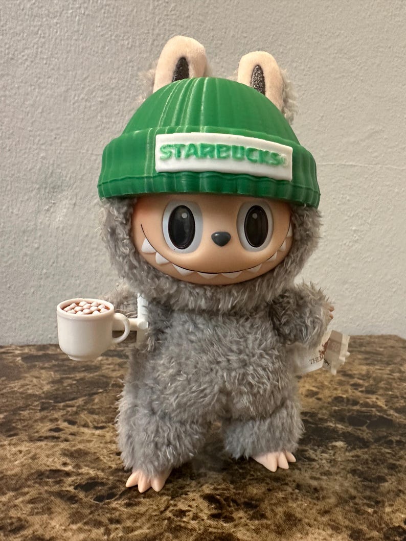 Puede incluir: Una figura de peluche de un personaje de dibujos animados que lleva un gorro verde con "STARBUCKS" impreso. La figura sostiene una peque&ntilde;a taza blanca con frijoles marrones y un papel peque&ntilde;o. La criatura tiene pelaje gris y una cara sonriente.