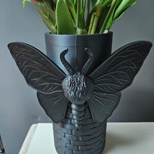 May include: Black decorative vase with a butterfly design. The vase has a cylindrical shape with a brick-like texture at the base and a butterfly with outstretched wings. The vase holds colorful artificial flowers.