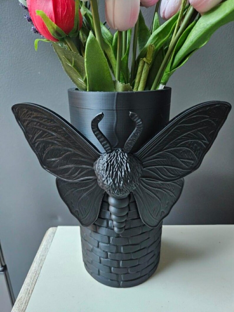 May include: Black decorative vase with a butterfly design. The vase has a cylindrical shape with a brick-like texture at the base and a butterfly with outstretched wings. The vase holds colorful artificial flowers.