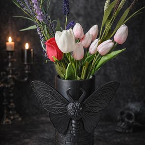 May include: A black vase with a moth design holds a bouquet of flowers, including pink and white tulips, a red rose, and purple lavender. The vase has a brick-like texture and is set against a dark background.
