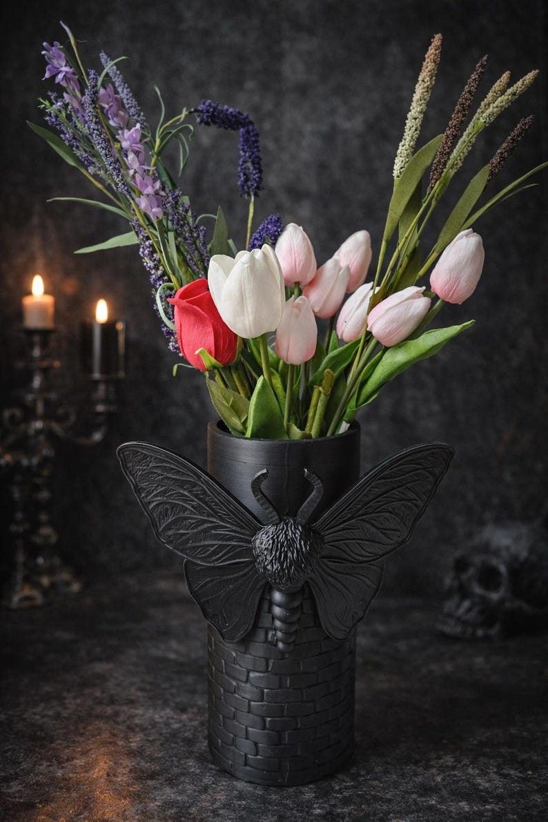 May include: A black vase with a moth design holds a bouquet of flowers, including pink and white tulips, a red rose, and purple lavender. The vase has a brick-like texture and is set against a dark background.
