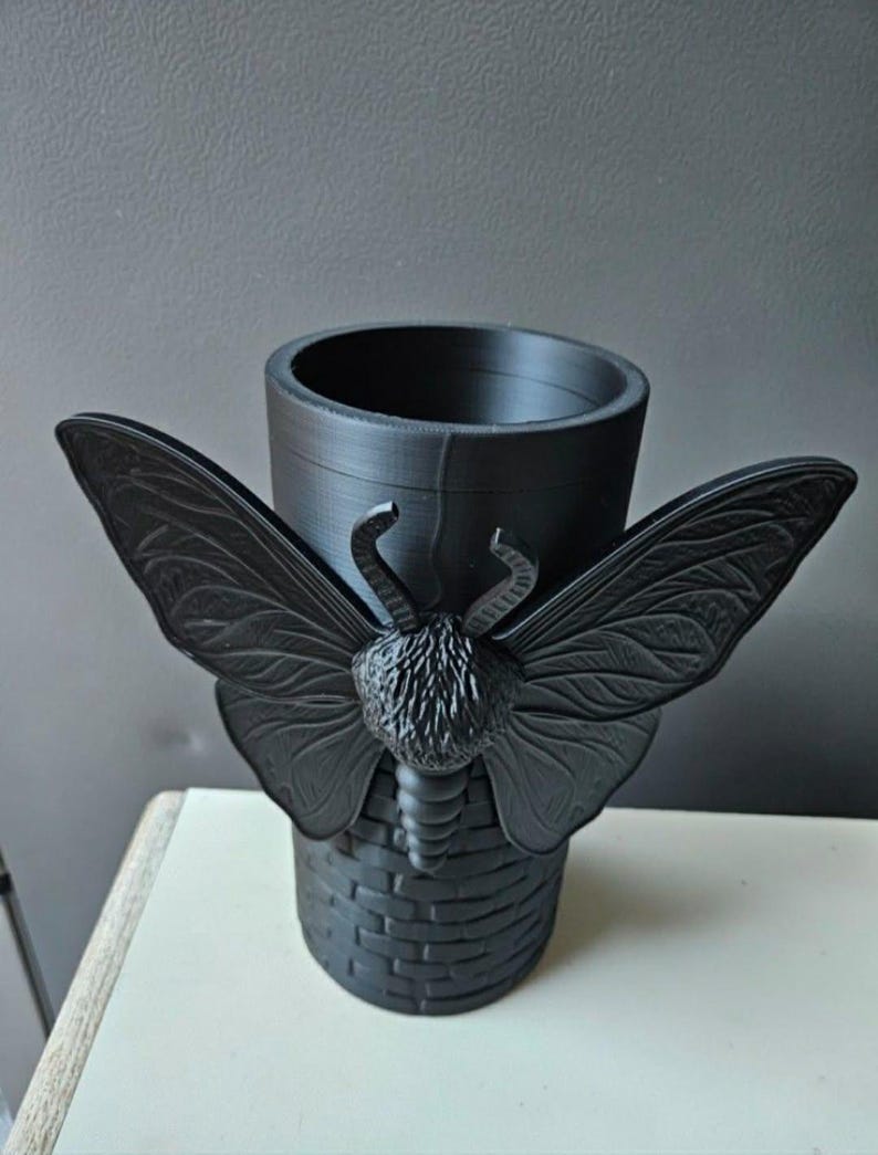 May include: A black decorative container with a moth design. The container has a cylindrical shape with a basket-weave base and a moth with outstretched wings. The moth has detailed wing patterns and a textured body.