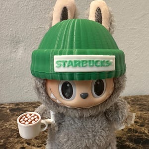 Puede incluir: Una figura de peluche de una criatura que lleva un gorro verde con el logo de Starbucks. La figura sostiene una peque&ntilde;a taza blanca llena de malvaviscos marrones. La figura tiene pelaje gris y ojos negros grandes.