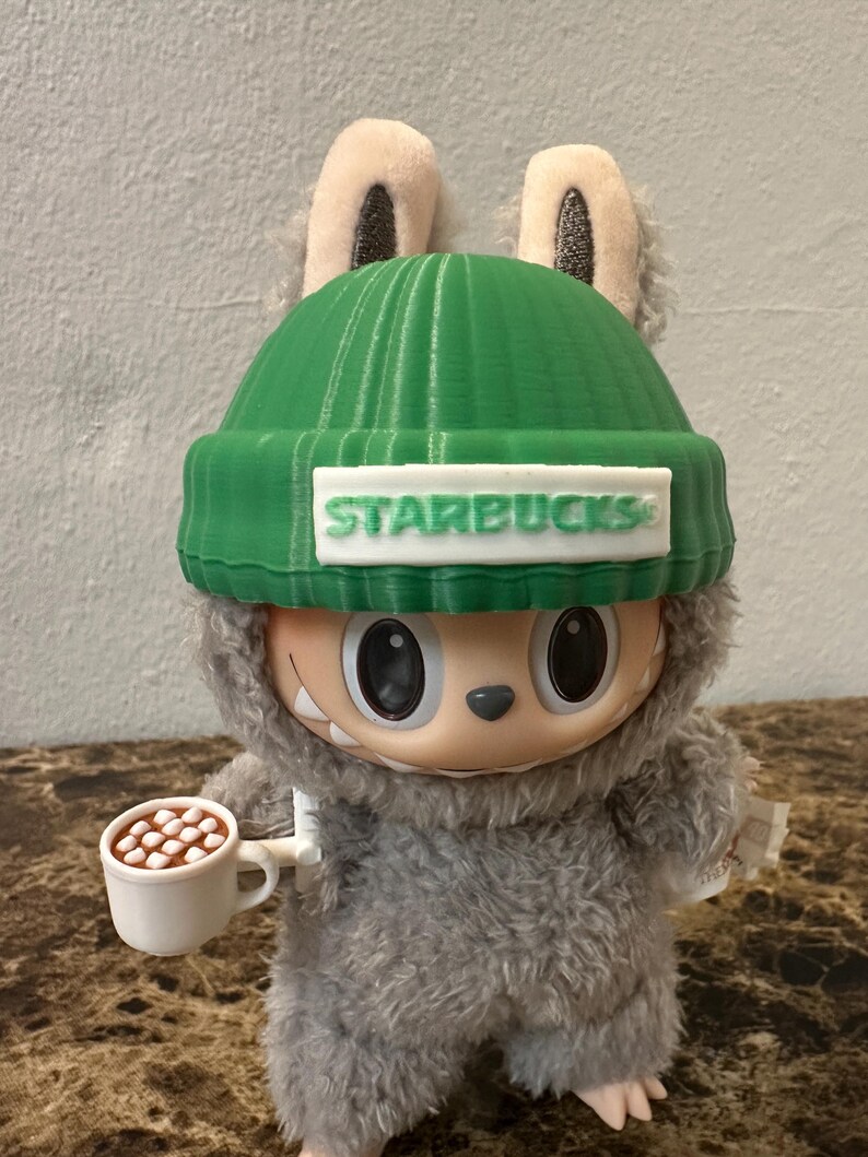 Puede incluir: Una figura de peluche de una criatura que lleva un gorro verde con el logo de Starbucks. La figura sostiene una peque&ntilde;a taza blanca llena de malvaviscos marrones. La figura tiene pelaje gris y ojos negros grandes.