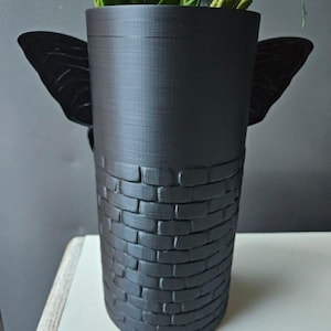 May include: A black decorative vase with a cylindrical shape. The top portion is smooth, while the bottom half features a brick pattern. Two black butterfly wings are attached to the sides. The vase holds green and yellow artificial plants.