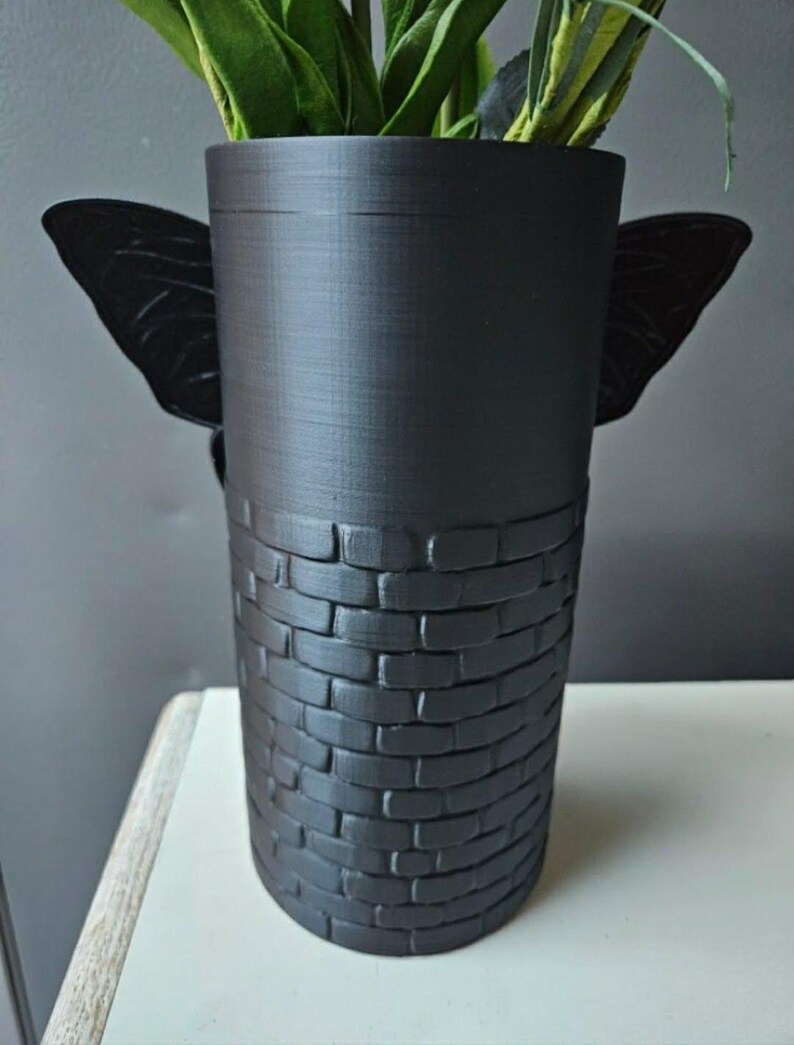 May include: A black decorative vase with a cylindrical shape. The top portion is smooth, while the bottom half features a brick pattern. Two black butterfly wings are attached to the sides. The vase holds green and yellow artificial plants.