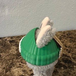 Puede incluir: Un peluche gris y blanco que lleva un caparaz&oacute;n verde brillante. El caparaz&oacute;n tiene una textura acanalada. El peluche tiene una etiqueta que dice "THE MONSTERS".