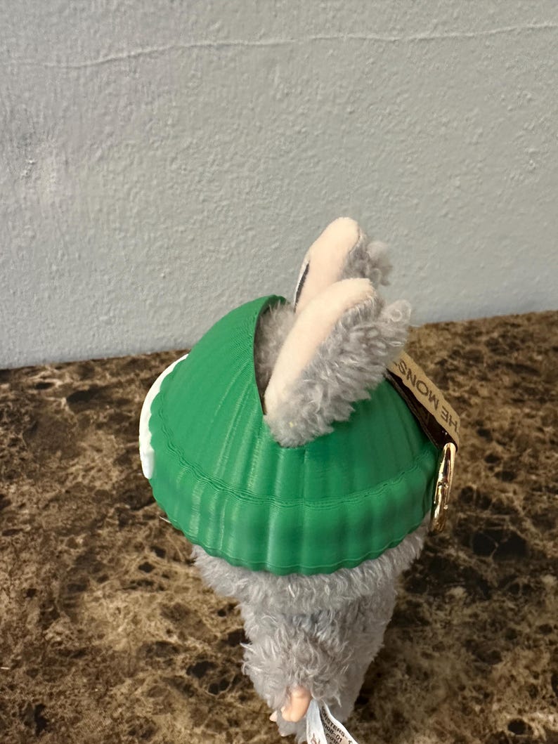 Puede incluir: Un peluche gris y blanco que lleva un caparaz&oacute;n verde brillante. El caparaz&oacute;n tiene una textura acanalada. El peluche tiene una etiqueta que dice "THE MONSTERS".