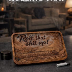 May include: A rectangular wooden rolling tray with the text "Roll that shit up!" and cannabis leaf designs. The tray is brown and has a raised edge. A black grinder and a silver vape pen are next to the tray. The words "PERSONALIZED ROLLING TRAY" are at the top.