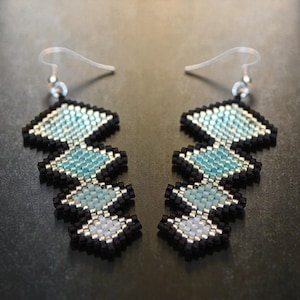 May include: A pair of handmade beaded earrings with a geometric design. The earrings feature a black border with light blue, silver, and white beads. The earrings hang from silver-colored ear wires.