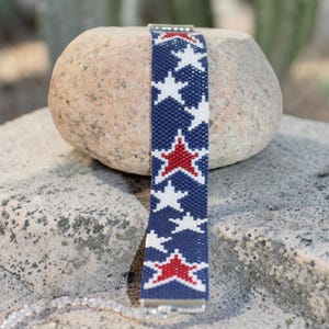 May include: A beaded bracelet with a patriotic design. The bracelet features a navy blue background with white and red star patterns. The bracelet is made of small beads and has a silver clasp.