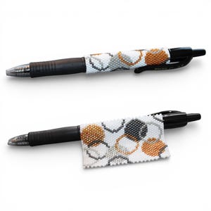 May include: Two black Pilot G-2 pens with beaded wraps. The wraps feature a pattern of white, gray, and gold-colored circles. The pens are shown against a white background.