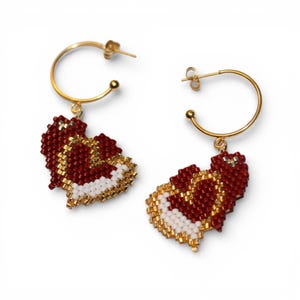 Puede incluir: Un par de pendientes de aro dorados con colgantes en forma de corazón. Los colgantes están hechos de pequeñas cuentas rojas, blancas y doradas, formando un diseño de corazón. Los pendientes se muestran sobre un fondo blanco.